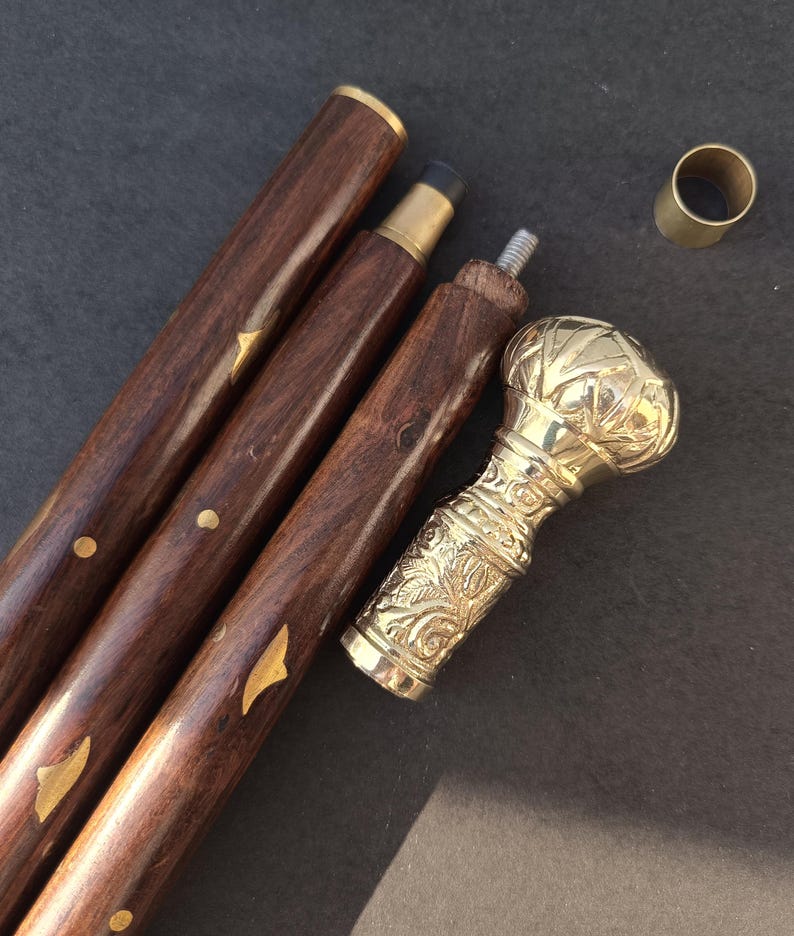 May include: A disassembled wooden walking cane with a brass handle and accents. The dark brown wooden shaft is in three sections, with gold-colored metal details. The handle is ornate, with a bulbous top and floral carvings.