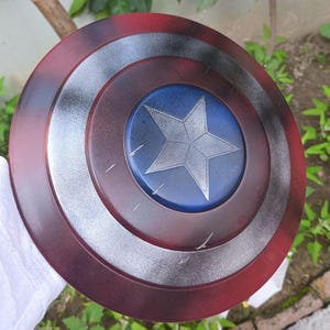 May include: A round shield with a red, silver, and blue color scheme. The shield features a central blue circle with a silver star, surrounded by concentric rings of silver and red. The shield is a prop or collectible.