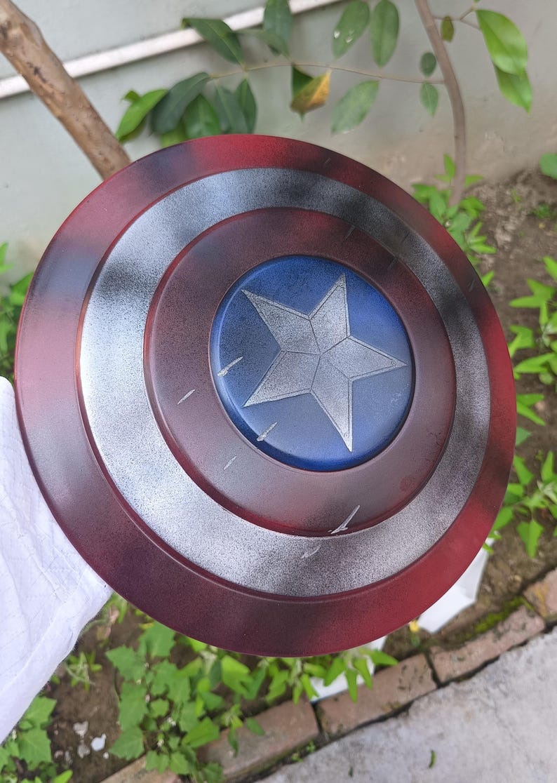 May include: A round shield with a red, silver, and blue color scheme. The shield features a central blue circle with a silver star, surrounded by concentric rings of silver and red. The shield is a prop or collectible.
