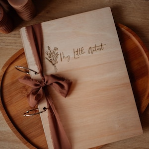 Op de afbeelding: Een houten schetsboek met de tekst "My Little Artist" en een bloemenillustratie. Het schetsboek is gebonden met zilveren ringen en een bruin lint. Het rust op een houten dienblad, met kaarsen op de achtergrond.