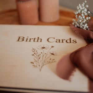 May include: A light-colored wooden book with the words "Birth Cards" engraved on the cover, along with a floral design. A brown ribbon and sprigs of white flowers are also visible.