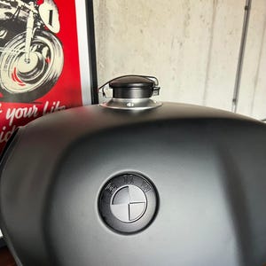 May include: A matte black motorcycle fuel tank featuring a centered black BMW emblem and a silver-toned metal fuel cap assembly with a black locking mechanism mounted on the top surface of the curved, rounded metal housing.