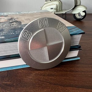Pode incluir: Um emblema circular da BMW em metal prateado com as letras icónicas da marca e um design circular segmentado. O emblema tem um acabamento mate escovado e está posicionado na vertical, exibindo claramente as letras B, M e W gravadas.
