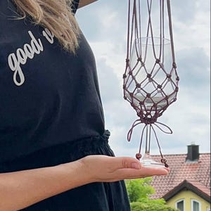 Könnte beinhalten: Ein handgefertigter Makramee-Pflanzenaufhänger in tiefem Burgunderrot, der ein klares Glas halten soll. Das Makramee hat ein geknotetes Netzdesign und wird an einer Schlaufe aufgehängt. Das Glas wird vom Makramee gehalten.