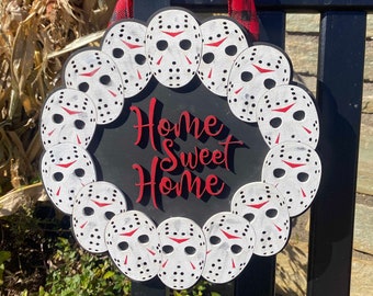 May include: A circular black wooden sign featuring the text Home Sweet Home in red script lettering, surrounded by a border of white hockey masks with red accents, suspended by a red and black buffalo plaid ribbon for hanging display.