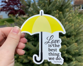 May include: A die-cut sticker featuring a bright yellow umbrella with a black handle and shaft. The text printed in black script and serif font reads: Love is the best thing we do. - Ted Mosby. The sticker has a white border and a matte finish.
