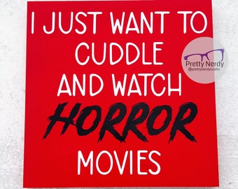 May include: A square red sign with white text that reads I JUST WANT TO CUDDLE AND WATCH, followed by the word HORROR in a jagged black font, and the word MOVIES in white. A circular logo with glasses and the text Pretty Nerdy is visible.