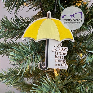 May include: Yellow umbrella ornament with the quote "Love is the best thing we do." - Ted Mosby. The ornament is hanging on a green Christmas tree.