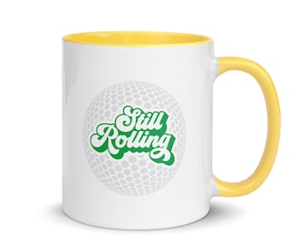 May include: White ceramic mug with a yellow rim and handle. The mug features a golf ball with the text "Still Rolling" in green.