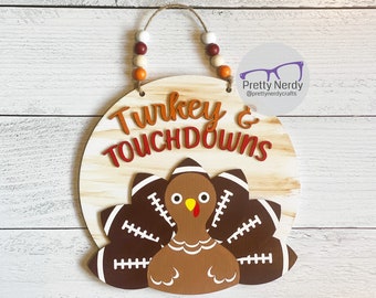 May include: A round wooden wall sign featuring the text "Turkey & TOUCHDOWNS" in orange and red. Below the text is a brown turkey with a yellow beak and red wattle, with its tail feathers depicted as brown footballs. It hangs from a jute rope.