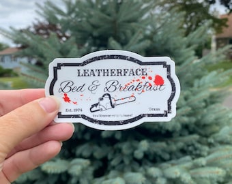 May include: A white rectangular sticker with a black border featuring the text LEATHERFACE Bed & Breakfast, Est. 1974, Texas, and You'll never want to leave! A black chainsaw illustration is centered with red paint splatters across the design.