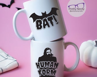 May include: Two white ceramic mugs with black graphic designs. The top mug has a bat silhouette with the word "BAT!" in a bold font. The bottom mug has a silhouette of a man with a beard and the words "HUMAN FORM" in a bold font.