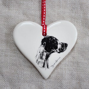 May include: A white, heart-shaped ceramic ornament with a black and white dog illustration. It has a red and white polka-dot ribbon for hanging. The dog portrait is signed with a cursive signature.