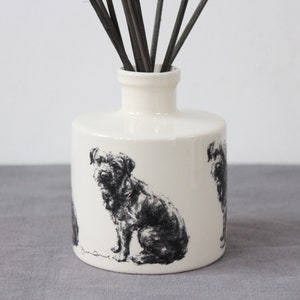 May include: White ceramic diffuser bottle with a black and white illustration of a dog. The bottle has a cylindrical shape and a narrow neck. Black diffuser sticks are inserted into the bottle.