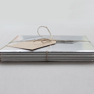 K&ouml;nnte beinhalten: Ein Stapel silberfarbenes Papier oder Karton, mit Bindfaden und einem kleinen braunen Etikett gebunden. Das Papier ist ordentlich gestapelt und verpackt, was auf ein Set von Schreibwaren oder Drucken hindeutet. Der Bindfaden ist zu einer einfachen Schleife gebunden.