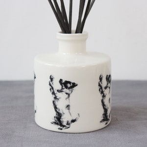 May include: White ceramic diffuser bottle with black and white illustrations of dogs. The bottle has a round shape and a narrow neck. It is filled with black diffuser sticks.