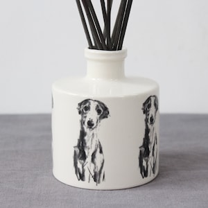 May include: White ceramic diffuser bottle with a black and white illustration of a Greyhound dog on the front. The bottle has a black reed diffuser stick inserted into the top.