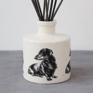 May include: White ceramic diffuser bottle with a black and white illustration of a dachshund. The bottle has black diffuser sticks.