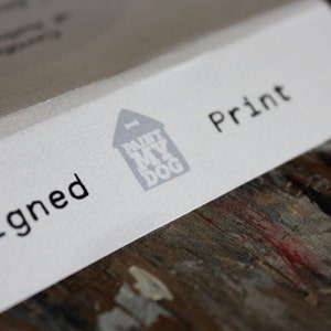 Op de afbeelding: Een witte kaart met de woorden "Gesigneerd" en "Print" in zwarte tekst. Een grijs huis met een hondenbot erbovenop en de woorden "Paint My Dog" erin, bevindt zich in het midden van de kaart.