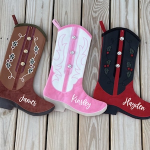 May include: Three cowboy boot-shaped Christmas stockings in brown, pink, and black. The brown boot has holly accents and the name "James". The pink boot has the name "Kinsley". The black boot has holly accents and the name "Hayden".