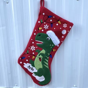 May include: Red Christmas stocking with a green dinosaur wearing a Santa hat. The stocking is decorated with white snowflakes, colorful lights, and the name "Arlo" in white script.