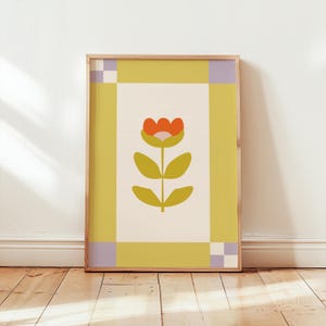Pode incluir: Uma impressão de arte emoldurada com uma flor estilizada com uma flor laranja e folhas verde-oliva. A obra de arte é definida contra um fundo creme, emoldurada por uma borda amarela com detalhes em xadrez lavanda e branco. A moldura é de madeira clara.