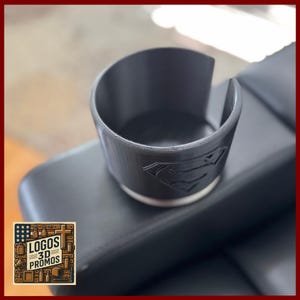 Puede incluir: Un portavasos negro con el logotipo de Superman en relieve en el lateral. El portavasos es cilíndrico con un recorte en un lado. Tiene un borde plateado en la base. El portavasos está sobre una superficie negra. La imagen también contiene el texto "LOGOS 3D PROMOS".