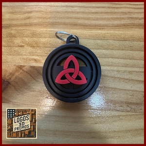 May include: Black and red keychain with a Celtic knot design. The keychain has a circular shape with a raised border and a metal ring for attachment. The Celtic knot is a three-part design.