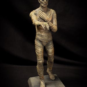 May include: A detailed figurine of a mummy, wrapped in aged bandages, stands on a rectangular base. The figure has a gaunt face and a green ring. The base is inscribed with "THE MUMMY".