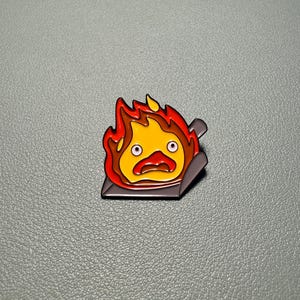 May include: A colorful enamel pin featuring a cartoon fire demon with a surprised expression. The pin has a yellow center, surrounded by orange and red flames, and is set on a dark gray base.