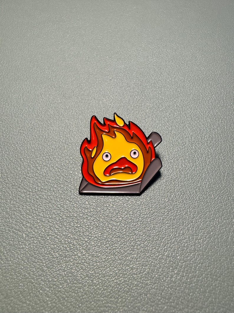 May include: A colorful enamel pin featuring a cartoon fire demon with a surprised expression. The pin has a yellow center, surrounded by orange and red flames, and is set on a dark gray base.