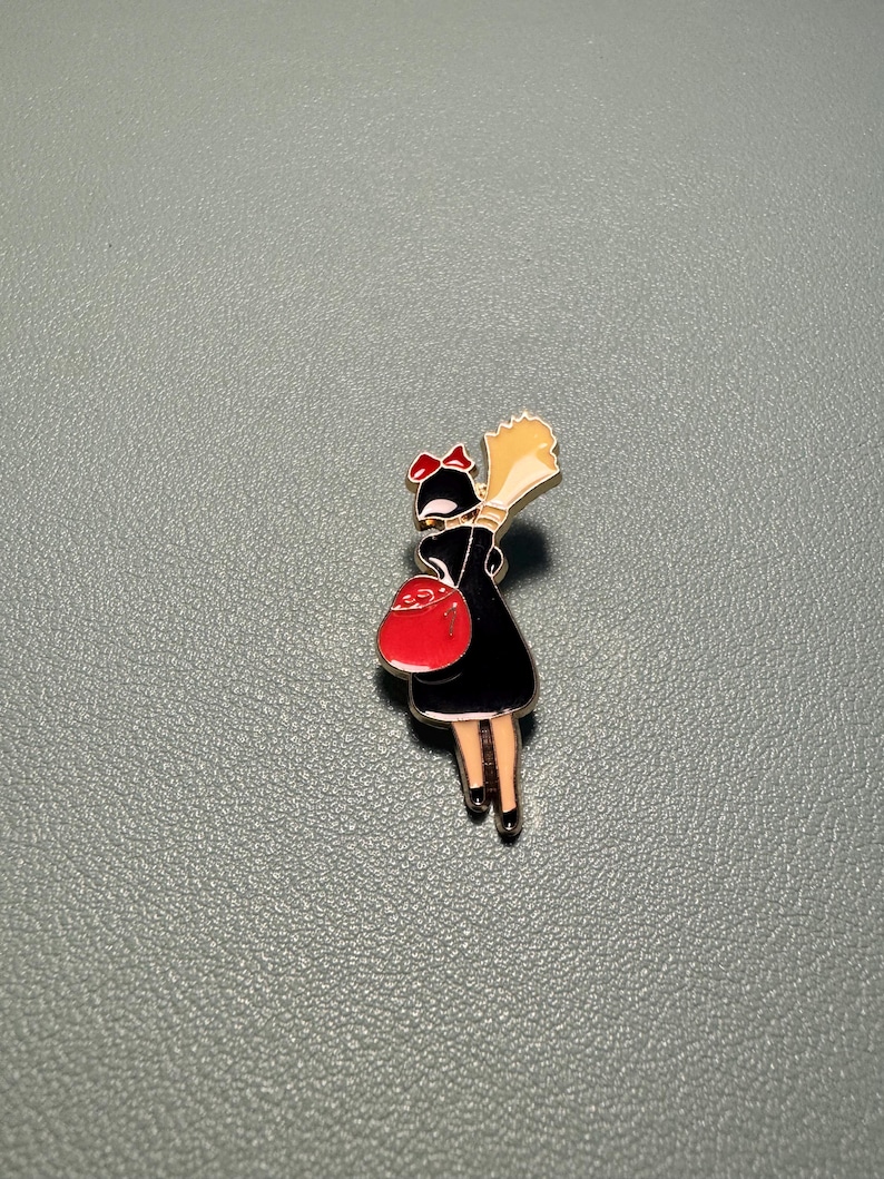 May include: Enamel pin featuring a cartoon character in a black dress, red bow, and red purse. The character is holding a bouquet of yellow flowers. The pin has a silver border and is set against a gray background.