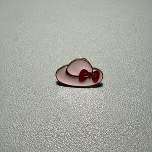 May include: A pink enamel hat pin with a red bow. The pin has a gold-colored outline and is set against a gray background. This accessory is a charming addition to any collection.