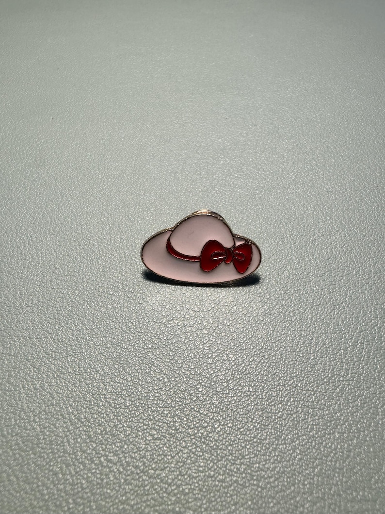 May include: A pink enamel hat pin with a red bow. The pin has a gold-colored outline and is set against a gray background. This accessory is a charming addition to any collection.
