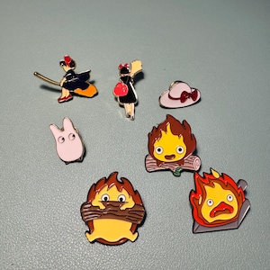 May include: A selection of enamel pins depicting characters and imagery from animated films. The pins include a witch on a broom, a girl with a red bag, a white hat with a red bow, a pink bunny, and several fire-themed characters.