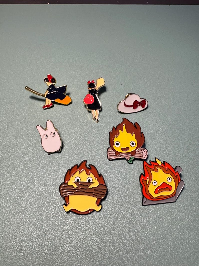 May include: A collection of enamel pins featuring characters and imagery from animated films. The pins include a witch on a broom, a girl with a red bag, a white hat with a red bow, a pink bunny, and several fire-themed characters.