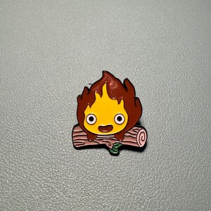May include: A colorful enamel pin featuring a cartoon fire demon with a yellow face, brown flames, and a smiling expression. The pin is set on a pink log with a green leaf.