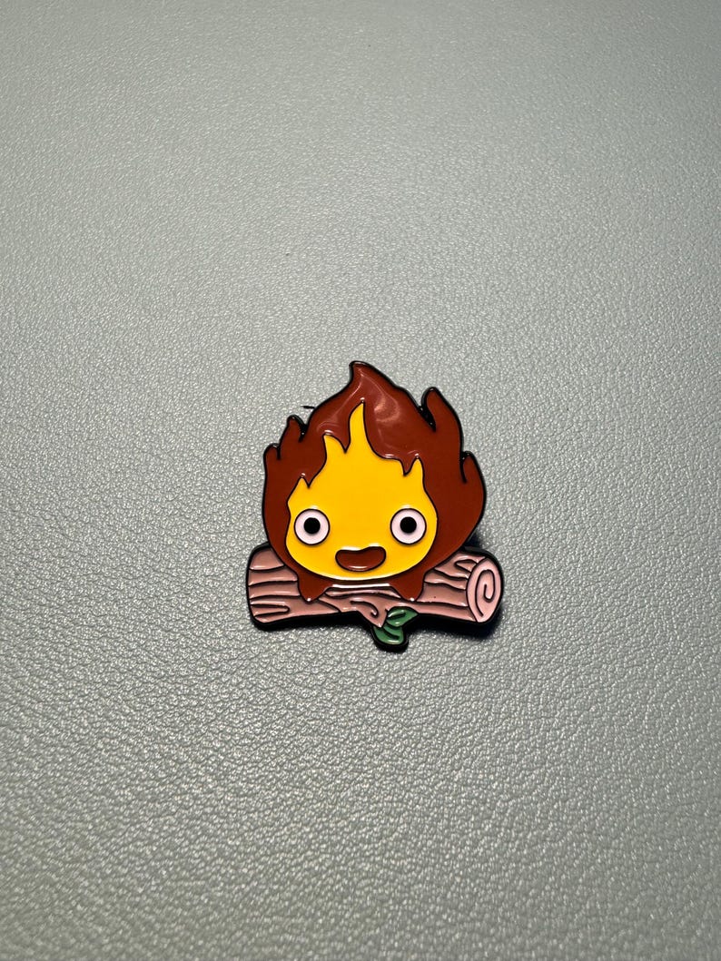 May include: A colorful enamel pin featuring a cartoon fire demon with a yellow face, brown flames, and a smiling expression. The pin is set on a pink log with a green leaf.