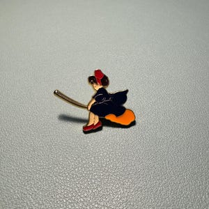 May include: Enamel pin depicting a witch riding a broom. The witch is wearing a black dress, red shoes, and a red hat. The broom is gold-colored, and the witch's skirt is orange. The pin is set against a gray background.