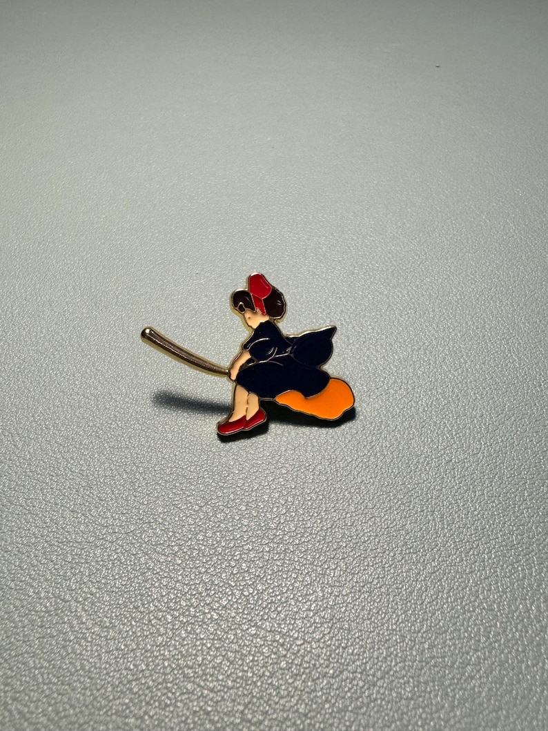 May include: Enamel pin depicting a witch riding a broom. The witch is wearing a black dress, red shoes, and a red hat. The broom is gold-colored, and the witch's skirt is orange. The pin is set against a gray background.