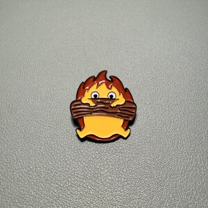 May include: A cartoon-style enamel pin featuring a yellow creature with brown eyes and a brown log in its mouth, surrounded by orange and red flames. The pin has a glossy finish and a black outline.