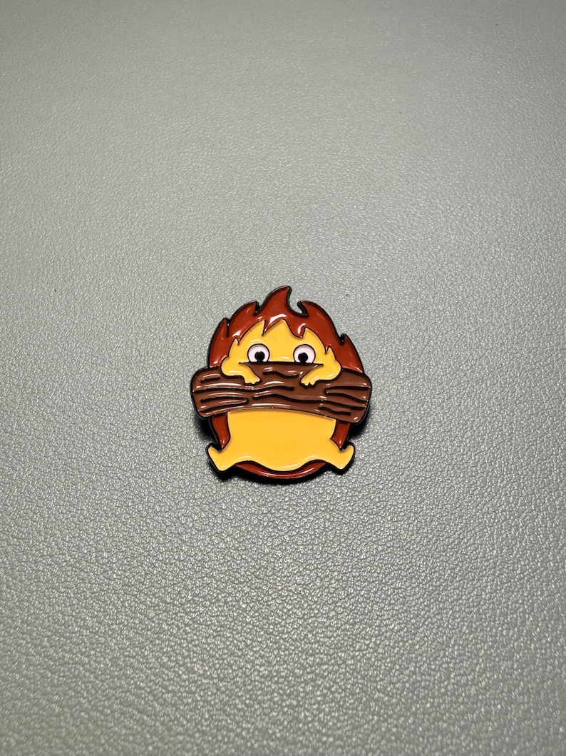 May include: A cartoon-style enamel pin featuring a yellow creature with brown eyes and a brown log in its mouth, surrounded by orange and red flames. The pin has a glossy finish and a black outline.