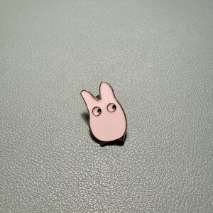 May include: A light pink enamel pin in the shape of a cartoon creature with two large eyes and small feet. The pin has a silver outline and is set against a gray background.