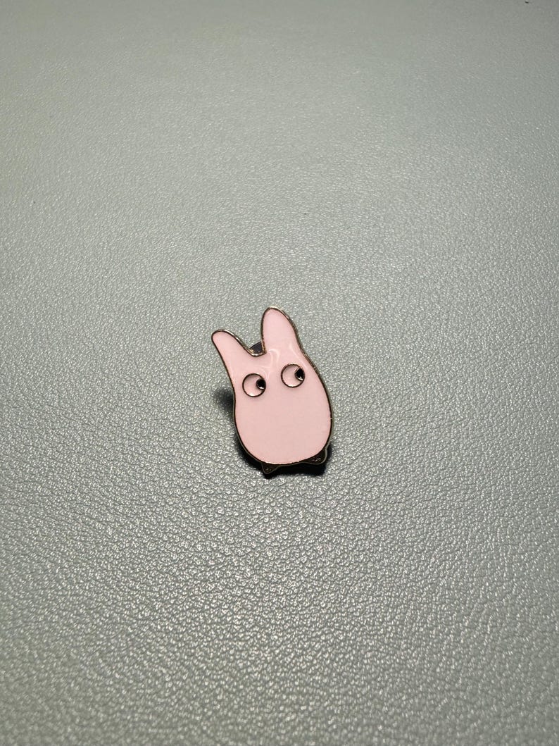 May include: A light pink enamel pin in the shape of a cartoon creature with two large eyes and small feet. The pin has a silver outline and is set against a gray background.