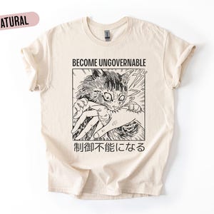 Puede incluir: Camiseta color crema con el texto "BECOME UNGOVERNABLE" sobre una ilustración en blanco y negro de estilo manga de un gato mordiendo una mano. Texto japonés debajo. La camiseta tiene mangas cortas y cuello redondo.