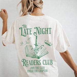 Può includere: T-shirt color crema con grafica verde. Il design presenta il testo "The Late Night Readers Club" con un'illustrazione di un libro, una spada e una corona. Sotto il testo ci sono le parole "Join the Club, Finish the Chapter".