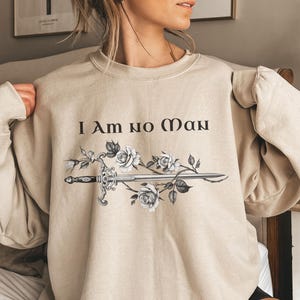 May include: Beige sweatshirt featuring the text "I Am no Man" in black, with a sword design and white roses. The long-sleeved crew neck top is made of a soft material.