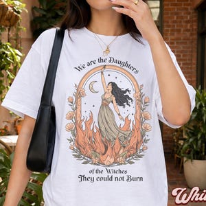 May include: White t-shirt featuring a graphic of a woman in a dress with flames, a crescent moon, and floral accents. The text reads "We are the Daughters of the Witches They could not Burn."
