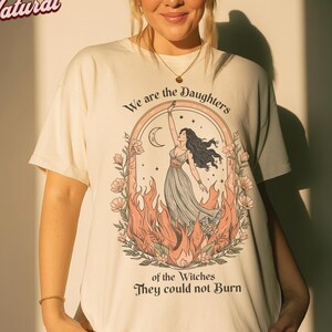 May include: Cream-colored t-shirt featuring a graphic of a woman with long black hair, reaching towards a crescent moon. The design is framed by flames and flowers, with the text "We are the Daughters of the Witches They could not Burn."