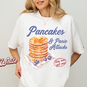 Peut inclure: T-shirt blanc avec l'inscription bleue "Pancakes & Panic Attacks". L'image présente une pile de crêpes avec du beurre et du sirop, ainsi que des myrtilles. Un graphique "achetez-en un, obtenez-en un" est également présent.